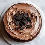 Chocolate cake oreo frosting - the image is of a round chocolate cake on a white marble countertop. the cake is covered in a thick layer of chocolate frosting, which is spread evenly over the top of the cake. on top, there are several oreo cookies scattered around the cake, adding a pop of color to the dish. the cookies are dark brown in color and appear to be freshly baked. the frosting is smooth and shiny, and the cake looks moist and fluffy.