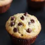 Chocolate chip muffin 4 ingredients - the image shows three freshly baked muffins on a black slate plate. the muffins are golden brown in color and have chocolate chips scattered on top. they are arranged in a row, with one muffin in the foreground and two in the background. the background is blurred, but it appears to be a kitchen countertop. the overall mood of the image is warm and inviting.