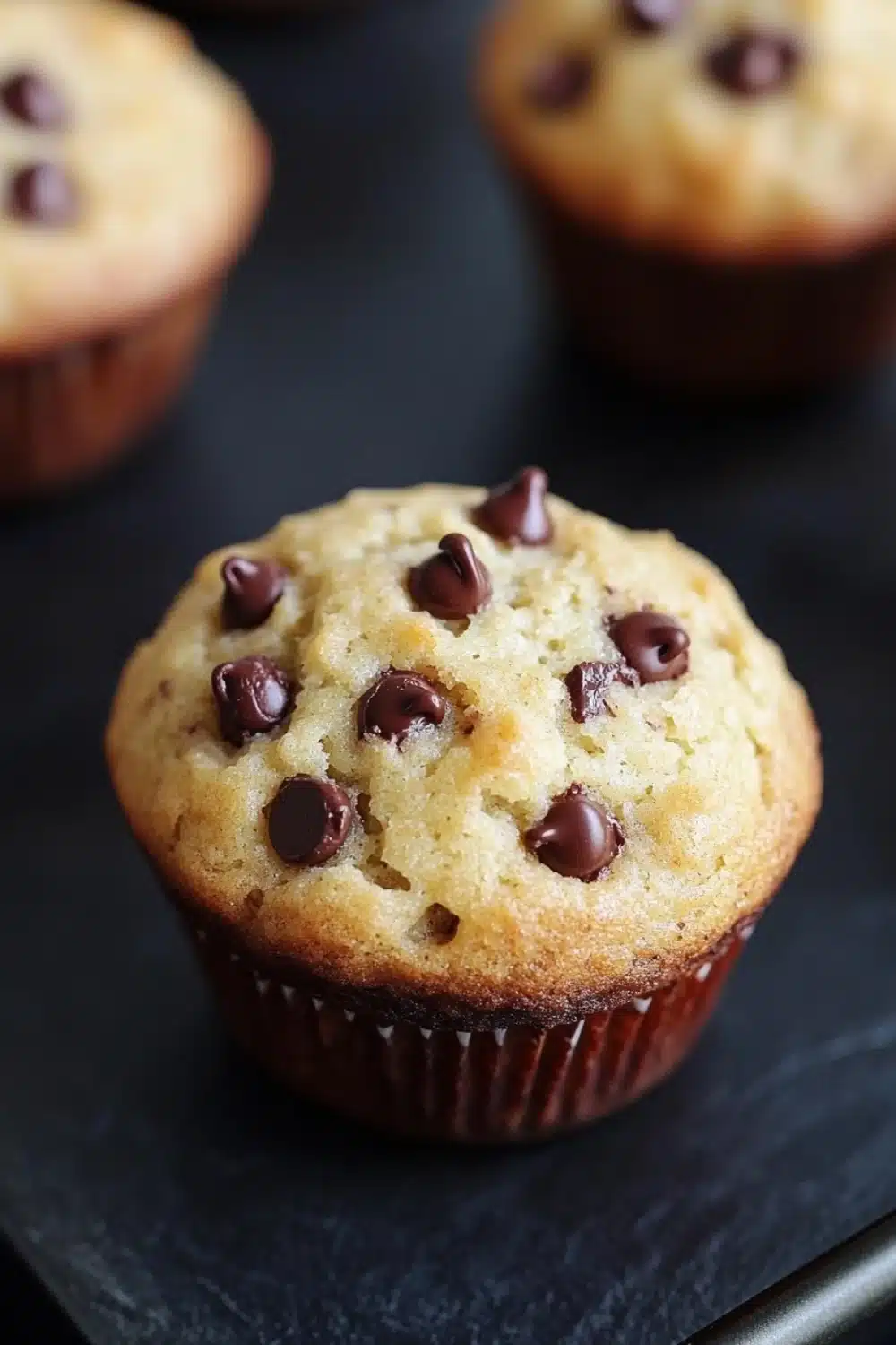 Chocolate chip muffin 4 ingredients - the image shows three freshly baked muffins on a black slate plate. the muffins are golden brown in color and have chocolate chips scattered on top. they are arranged in a row, with one muffin in the foreground and two in the background. the background is blurred, but it appears to be a kitchen countertop. the overall mood of the image is warm and inviting.