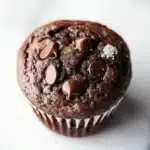 Chocolate muffin cup - the image is a close-up of a chocolate muffin on a white marble surface. the muffin is round and has a golden brown color. it is topped with chocolate chips and sprinkled with powdered sugar. the background is blurred, but it appears to be a kitchen countertop with other muffins visible in the top right corner.