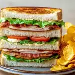 Club sandwich with chips - the image shows a sandwich on a plate with a side of potato chips. the sandwich is made with two slices of toasted bread, filled with ham, lettuce, tomato, and cheese. the bread is golden brown and appears to be crispy on the outside. the plate is blue and the chips are golden brown. the background is blurred, but it seems to be a wooden table.