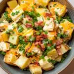 Creamy loaded baked potato salad - the image is a close-up of a bowl of potato salad. the bowl is made of light-colored wood and has a dark brown rim. the salad is made up of diced potatoes, diced green onions, bacon bits, and melted cheese. the cheese is a bright yellow color and appears to be melted and bubbly. the bacon bits are scattered throughout the salad, adding a pop of color and texture. the green onions are bright green and look fresh and vibrant. the overall appearance of the salad is fresh and appetizing.