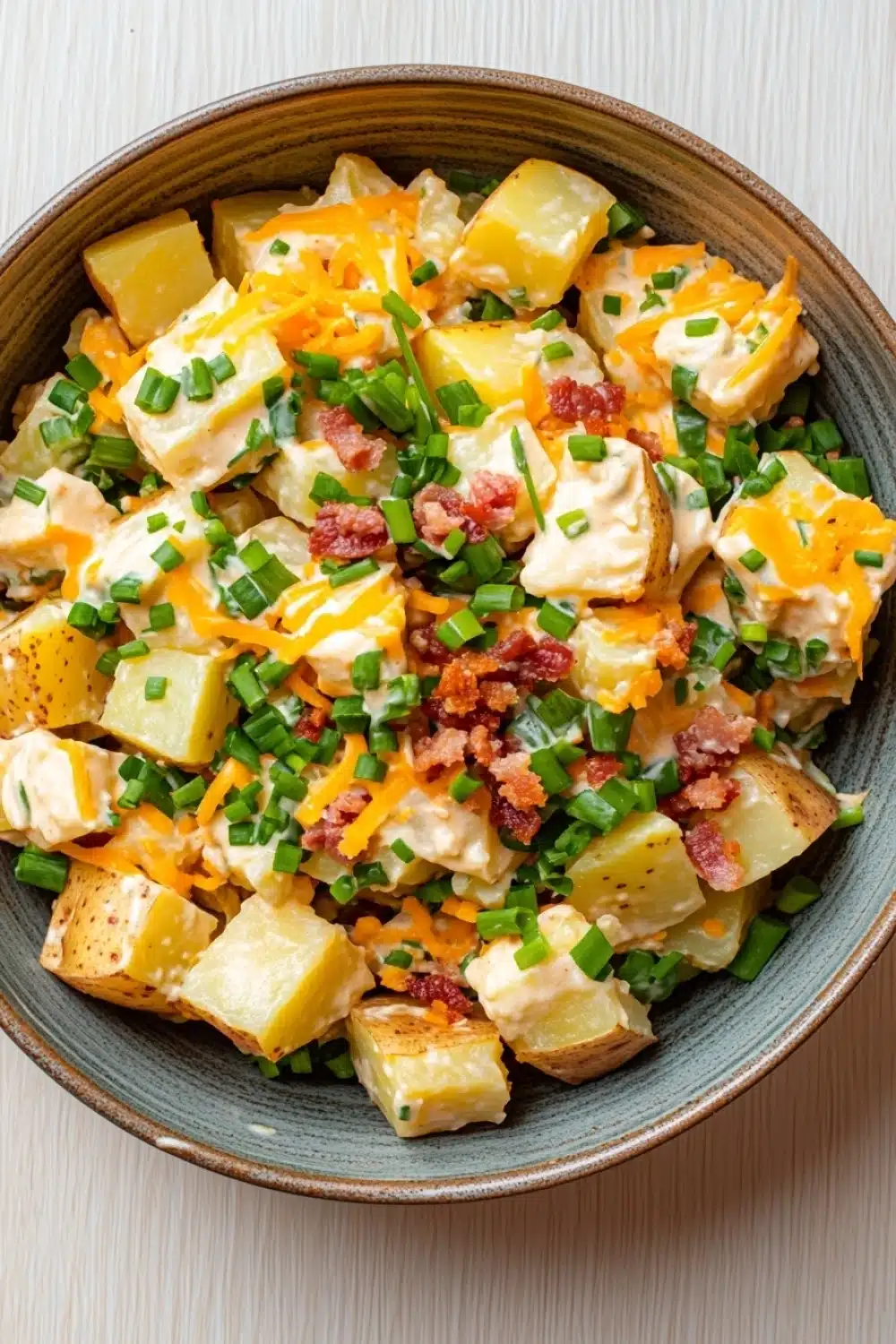 Creamy loaded baked potato salad - the image is a close-up of a bowl of potato salad. the bowl is made of light-colored wood and has a dark brown rim. the salad is made up of diced potatoes, diced green onions, bacon bits, and melted cheese. the cheese is a bright yellow color and appears to be melted and bubbly. the bacon bits are scattered throughout the salad, adding a pop of color and texture. the green onions are bright green and look fresh and vibrant. the overall appearance of the salad is fresh and appetizing.