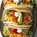 Crock pot chicken tacos - the image shows a plate with three tacos on it. the tacos are made with shredded chicken, shredded cheese, diced tomatoes, avocado, and a dollop of sour cream. the plate is brown and has a floral design around the edges. the background is a white marble countertop.