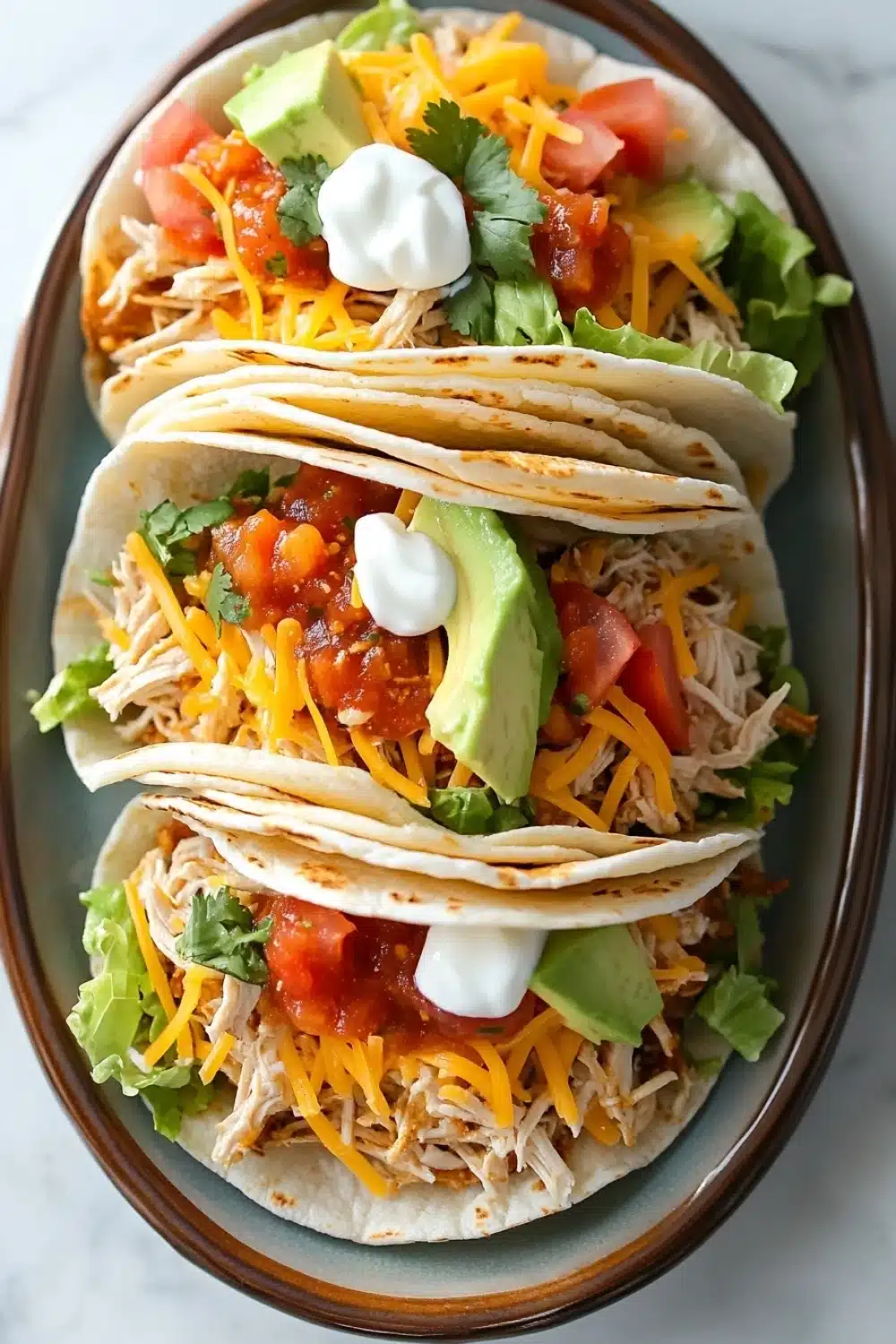 Crock pot chicken tacos - the image shows a plate with three tacos on it. the tacos are made with shredded chicken, shredded cheese, diced tomatoes, avocado, and a dollop of sour cream. the plate is brown and has a floral design around the edges. the background is a white marble countertop.
