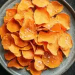 Dehydrated sweet potato chips air fryer - the image shows a plate of sweet potato chips on a dark wooden table. the chips are arranged in a circular pattern on the plate, with some overlapping each other. they are orange in color and have small white specks scattered throughout. the plate is made of a light-colored ceramic or ceramic material and has a textured surface. the background is blurred, but it appears to be a rustic wooden surface.