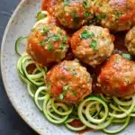 Dinner turkey meatballs with zucchini noodles - the image shows a plate of meatballs and zucchini noodles. the meatballs are round and appear to be seasoned with herbs and spices. the noodles are thin and green, and they are arranged in a circular pattern around the meatballs. the dish is garnished with chopped parsley. the plate is white with a speckled pattern, and the background is a dark grey countertop.