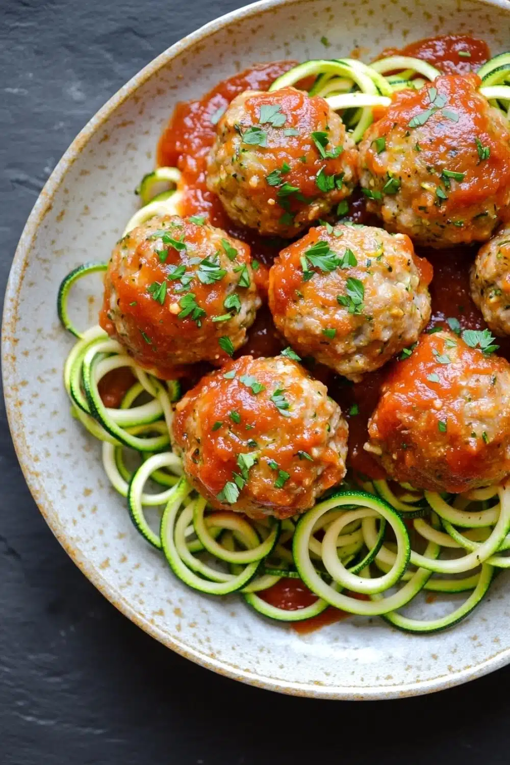 Dinner turkey meatballs with zucchini noodles - the image shows a plate of meatballs and zucchini noodles. the meatballs are round and appear to be seasoned with herbs and spices. the noodles are thin and green, and they are arranged in a circular pattern around the meatballs. the dish is garnished with chopped parsley. the plate is white with a speckled pattern, and the background is a dark grey countertop.