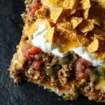 Dorito casserole freezer meal - the image shows a slice of nachos on a black slate plate. the nacho is topped with a layer of ground beef, diced tomatoes, green bell peppers, and melted cheese. the cheese is melted and bubbly, and there is a dollop of sour cream on top. there are also a few tortilla chips scattered around the plate.