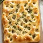 Focaccia bread - the image shows a square-shaped flatbread on a wooden cutting board. the flatbread appears to be freshly baked and has a golden-brown crust. it is topped with a generous amount of rosemary sprigs and sprinkled with sesame seeds. the cutting board is placed on a white wooden table with a gray cloth napkin next to it. the background is blurred, but it seems to be a kitchen countertop.