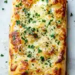 Garlic bread with pizza dough - the image shows a rectangular piece of bread on a white marble countertop. the bread appears to be freshly baked and has a golden brown crust. on top of the bread, there is a layer of melted cheese and a sprinkle of chopped parsley. the cheese is melted and bubbly, and there are small pieces of bread crumbs scattered around the edges. the parsley is bright green and looks fresh and vibrant. the background is blurred, making the bread the focal point of the image.