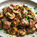 Garlic butter steak bites with mushrooms crockpot - the image is a close-up of a plate of food. the plate is white with a speckled pattern and is placed on a white marble countertop. the food appears to be a dish made with chunks of beef and mushrooms. the beef is cooked medium-rare and has a pinkish-brown color. the mushrooms are sliced and arranged in a pile on top of the beef. there are also some chopped parsley scattered around the plate, adding a pop of green color to the dish. the overall appearance of the dish is savory and appetizing.