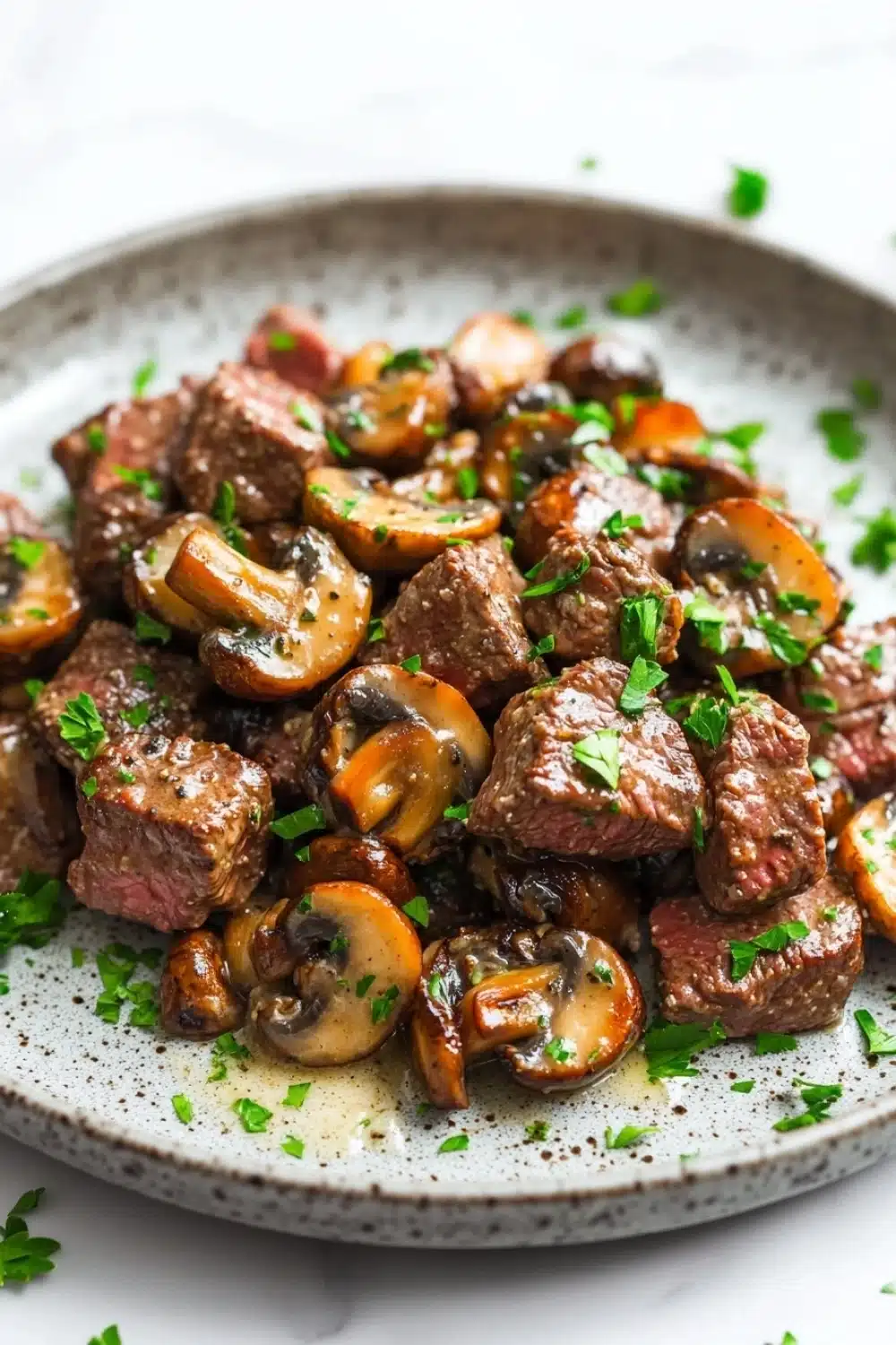 Garlic butter steak bites with mushrooms crockpot - the image is a close-up of a plate of food. the plate is white with a speckled pattern and is placed on a white marble countertop. the food appears to be a dish made with chunks of beef and mushrooms. the beef is cooked medium-rare and has a pinkish-brown color. the mushrooms are sliced and arranged in a pile on top of the beef. there are also some chopped parsley scattered around the plate, adding a pop of green color to the dish. the overall appearance of the dish is savory and appetizing.