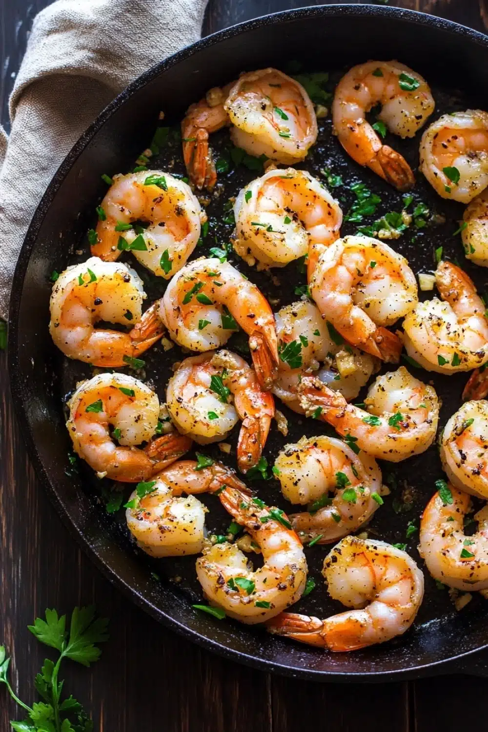 Garlic shrimp in cast iron skillet - the image shows a black cast iron skillet filled with cooked shrimp. the shrimp are orange in color and appear to be seasoned with herbs and spices. the skillet is sitting on a wooden table with a beige cloth napkin on the side. there are a few sprigs of parsley scattered around the skillet. the overall appearance of the dish is appetizing and ready to eat.