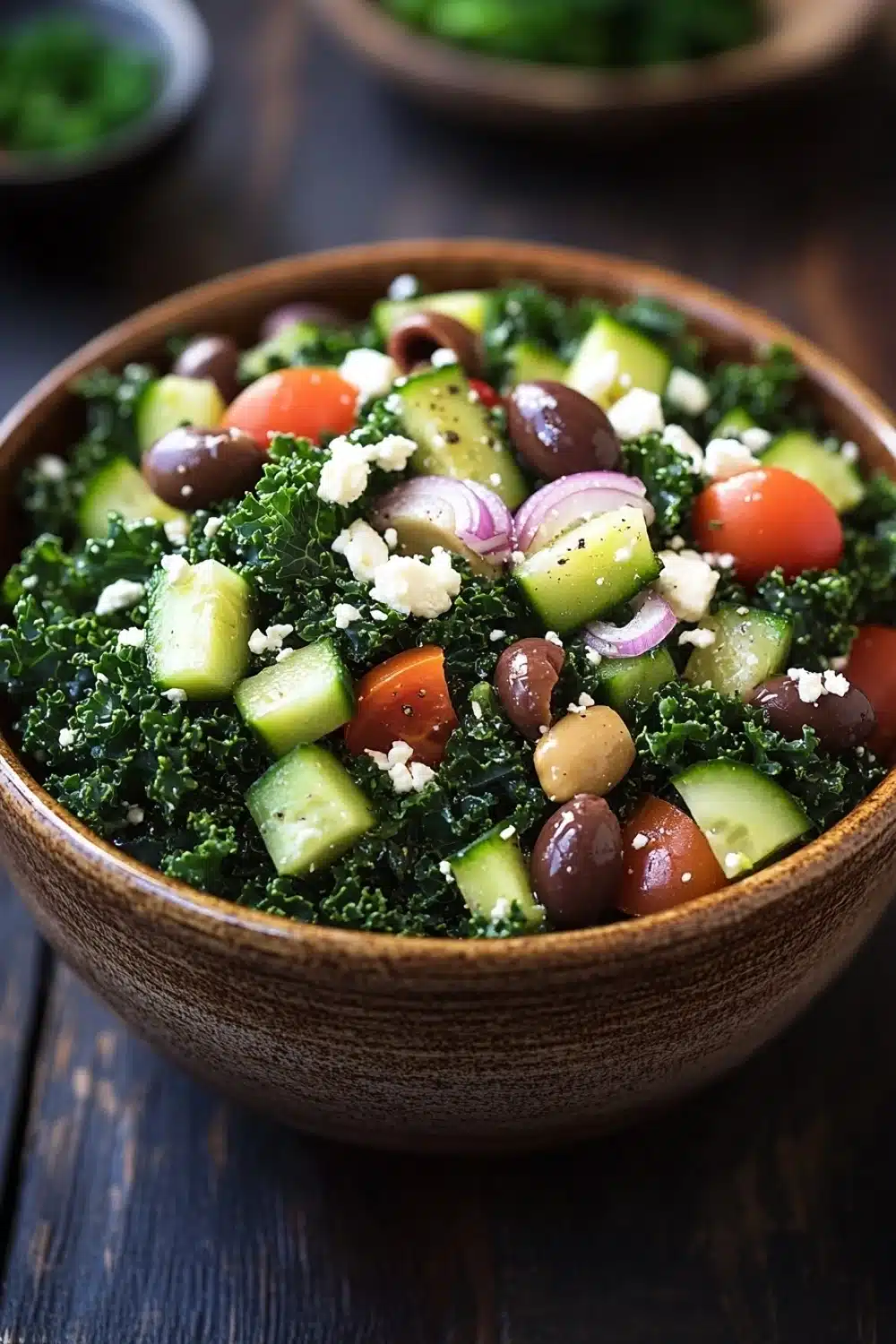 Greek salad kale - the image is a close-up of a wooden bowl filled with a colorful salad. the salad is made up of kale, sliced cucumbers, red onions, cherry tomatoes, and black olives. the kale is a vibrant green color and appears to be seasoned with herbs and spices. the olives are scattered throughout the salad, adding a pop of color and texture. the bowl is sitting on a dark wooden table, and there are other bowls of salad in the background. the overall mood of the image is fresh and appetizing.