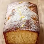 Greek yogurt lemon loaf cake - the image is a close-up of a freshly baked loaf of bread on a wooden cutting board. the bread is golden brown in color and has a crumbly texture. it is dusted with a light dusting of powdered sugar, giving it a shiny appearance. on top of the bread, there are small pieces of lemon zest scattered around, adding a pop of color to the dish. the background is blurred, making the bread the focal point of the image.