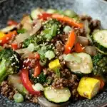 Ground beef and veggies - the image is a close-up of a plate of food. the plate is black and has a textured surface. the food appears to be a stir-fry dish with ground beef, sliced vegetables, and sesame seeds. the vegetables include broccoli, red bell peppers, zucchini, and yellow squash. there are also sliced onions and green onions scattered throughout the dish. the dish looks fresh and appetizing.
