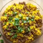 Ground beef and yellow rice for dinner - the image shows a large glass bowl filled with a mixture of yellow rice and ground meat. the rice appears to be cooked and has a golden brown color. the ground meat is scattered throughout the mixture, with some pieces of ground beef visible. the bowl is sitting on a wooden surface, and there are a few sprigs of cilantro scattered around it. the overall appearance of the dish is messy and appetizing.