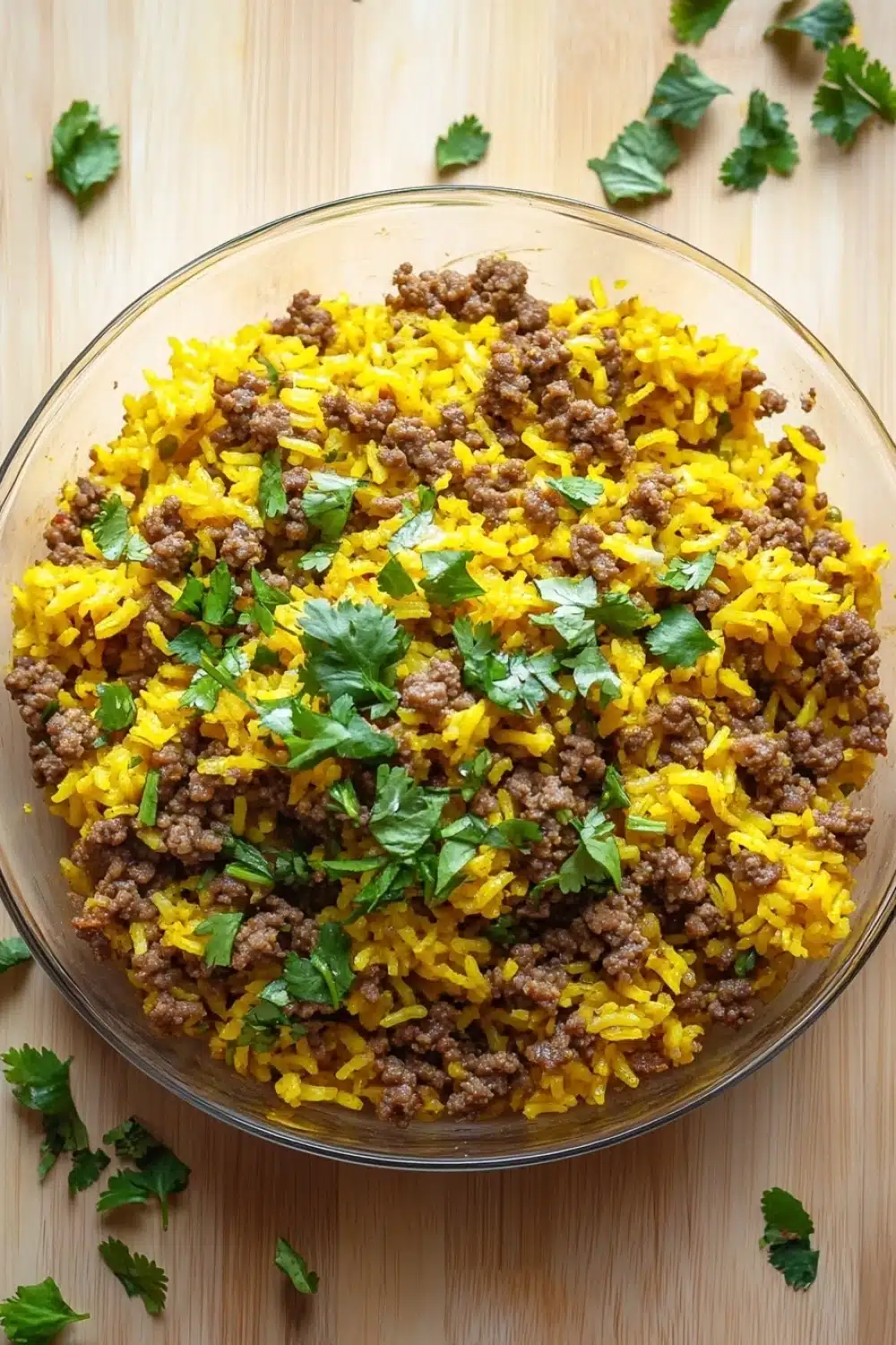 Ground beef and yellow rice for dinner - the image shows a large glass bowl filled with a mixture of yellow rice and ground meat. the rice appears to be cooked and has a golden brown color. the ground meat is scattered throughout the mixture, with some pieces of ground beef visible. the bowl is sitting on a wooden surface, and there are a few sprigs of cilantro scattered around it. the overall appearance of the dish is messy and appetizing.
