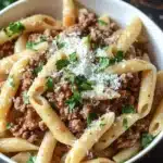 Ground beef pasta dish - the image shows a bowl of penne pasta with ground beef and parmesan cheese. the pasta is cooked al dente and is topped with a generous amount of minced meat. the dish is garnished with fresh parsley leaves. the bowl is white and is sitting on a wooden table. the background is blurred, but it appears to be a rustic kitchen countertop.