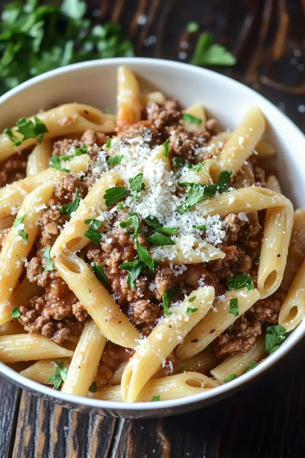 Ground beef pasta dish - the image shows a bowl of penne pasta with ground beef and parmesan cheese. the pasta is cooked al dente and is topped with a generous amount of minced meat. the dish is garnished with fresh parsley leaves. the bowl is white and is sitting on a wooden table. the background is blurred, but it appears to be a rustic kitchen countertop.