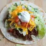 Ground beef skillet taco - the image is a close-up of a taco on a wooden table. the taco is made with a tortilla shell and is filled with ground beef, diced tomatoes, shredded cheese, and a dollop of sour cream on top. there are also two lime wedges on the side of the tortilla. the toppings are arranged in a colorful and appetizing manner. the background is blurred, but it appears to be a kitchen countertop.