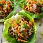 High protein beef lettuce wraps - the image shows four lettuce wraps filled with ground beef and vegetables. the lettuce wraps are arranged in a row on a wooden surface. the ground beef appears to be cooked and is mixed with diced carrots, onions, and green onions. the vegetables are bright orange and appear to be seasoned with herbs and spices. there are also some chopped green onions scattered around the lettuce wraps. the overall appearance of the wraps is fresh and appetizing.