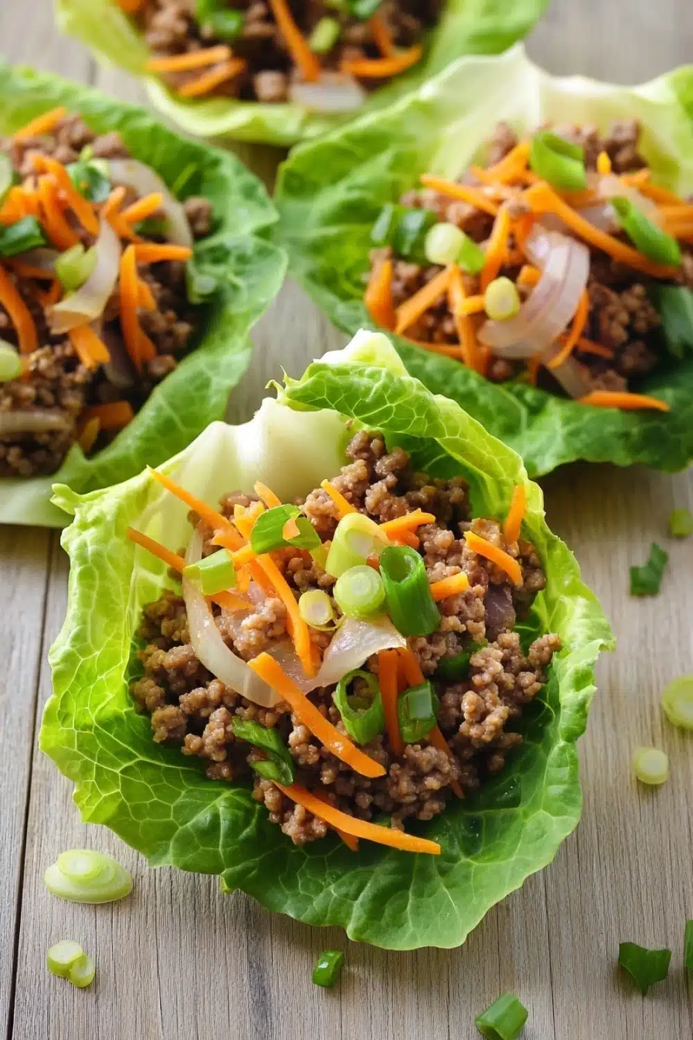 High protein beef lettuce wraps - the image shows four lettuce wraps filled with ground beef and vegetables. the lettuce wraps are arranged in a row on a wooden surface. the ground beef appears to be cooked and is mixed with diced carrots, onions, and green onions. the vegetables are bright orange and appear to be seasoned with herbs and spices. there are also some chopped green onions scattered around the lettuce wraps. the overall appearance of the wraps is fresh and appetizing.