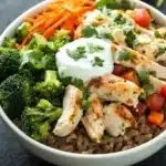 High protein chicken and rice bowl - the image is a close-up of a bowl of food. the bowl is white and is placed on a black countertop. the food in the bowl appears to be a stir-fry dish with chicken, broccoli, carrots, and other vegetables. the chicken is cooked to a golden brown color and is garnished with fresh cilantro. there is also a dollop of sour cream on top of the chicken. the vegetables are bright red and appear to be diced tomatoes or bell peppers. the dish looks fresh and appetizing.