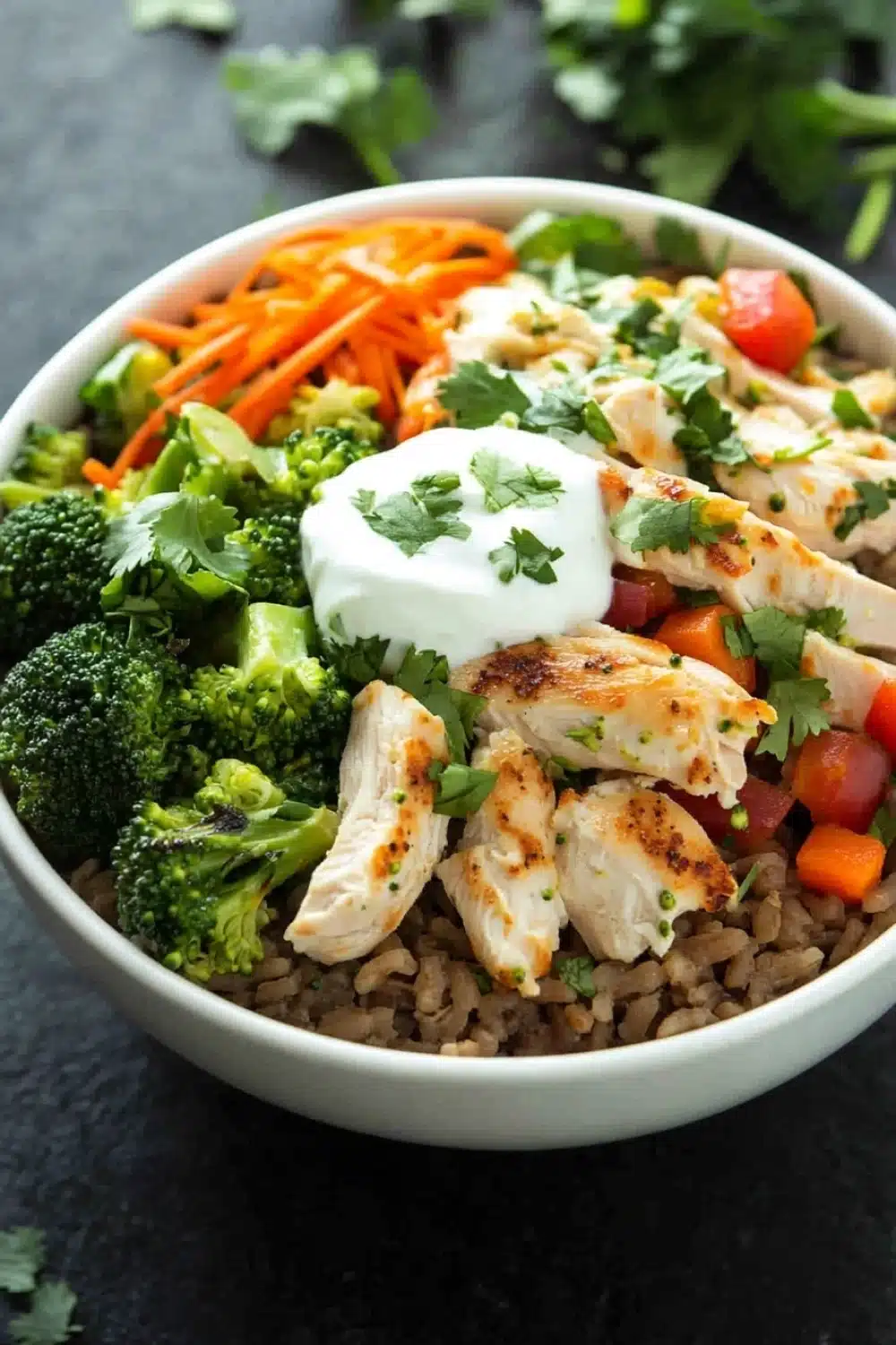 High protein chicken and rice bowl - the image is a close-up of a bowl of food. the bowl is white and is placed on a black countertop. the food in the bowl appears to be a stir-fry dish with chicken, broccoli, carrots, and other vegetables. the chicken is cooked to a golden brown color and is garnished with fresh cilantro. there is also a dollop of sour cream on top of the chicken. the vegetables are bright red and appear to be diced tomatoes or bell peppers. the dish looks fresh and appetizing.