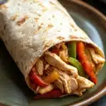 High protein chicken fajita burritos - the image shows a wrap on a green plate. the wrap is made of tortilla and is filled with shredded chicken, red and green bell peppers, and other vegetables. the chicken appears to be cooked and has a golden brown color. the vegetables are cut into small pieces and are arranged in a circular pattern around the wrap. the plate is sitting on a black countertop with a striped napkin in the background.