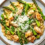 High protein chicken orzo with asparagus - the image is a close-up of a plate of pasta dish. the pasta is fusilli, which is a type of spiral-shaped pasta, and is topped with grilled chicken and asparagus. the chicken is cooked to a golden brown color and appears to be seasoned with herbs and spices. the dish is garnished with a sprinkle of parmesan cheese. the plate is sitting on a white marble countertop with a few sprigs of parsley scattered around.