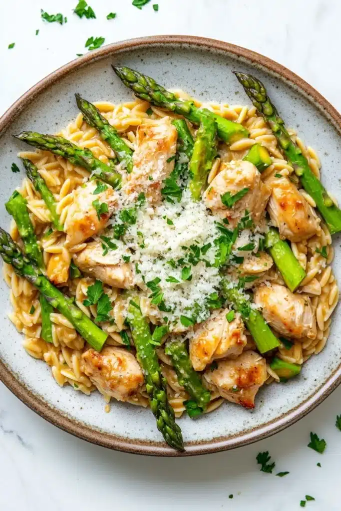 High protein chicken orzo with asparagus - the image is a close-up of a plate of pasta dish. the pasta is fusilli, which is a type of spiral-shaped pasta, and is topped with grilled chicken and asparagus. the chicken is cooked to a golden brown color and appears to be seasoned with herbs and spices. the dish is garnished with a sprinkle of parmesan cheese. the plate is sitting on a white marble countertop with a few sprigs of parsley scattered around.