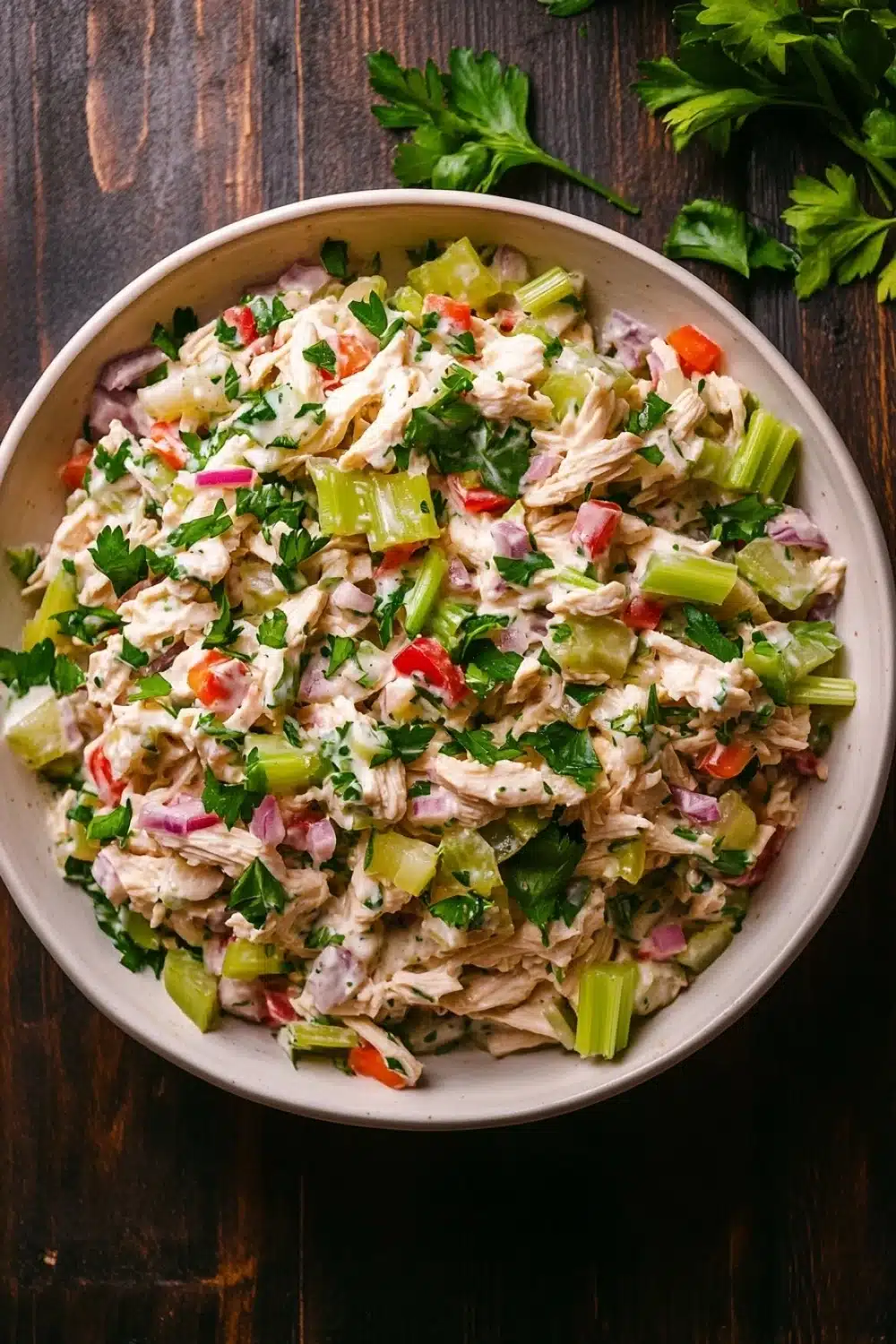 High protein chicken salad with greek yogurt - the image is a close-up of a large white bowl filled with a colorful salad. the salad appears to be made with shredded chicken, celery, red bell peppers, and other vegetables. the bowl is placed on a wooden table with a sprig of fresh cilantro on the side. the background is blurred, but it seems to be a rustic kitchen countertop. the overall color palette of the image is warm and inviting.