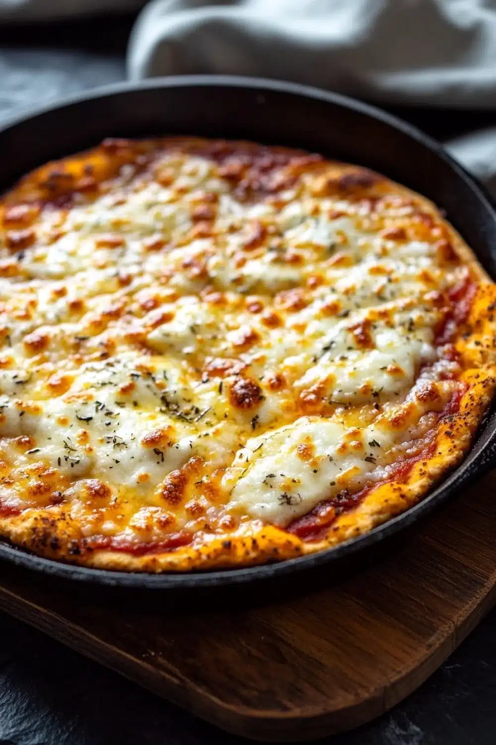 Keto pizza crust in cast iron pan - the image shows a freshly baked pizza in a black cast iron skillet on a wooden cutting board. the pizza has a golden brown crust and is topped with melted cheese and herbs. the cheese appears to be melted and bubbly, and there are small bits of red sauce scattered throughout the pizza. the background is blurred, but it seems to be a kitchen countertop with a white cloth napkin on the side.