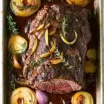 Keto slow cooker roast beef - the image shows a rectangular baking dish filled with a cooked steak. the steak appears to be medium-rare and has a dark brown crust on top. it is garnished with sliced onions and a sprig of thyme. the dish is sitting on a white marble countertop. there are also a few red onions scattered around the dish. the overall appearance of the dish is savory and appetizing.