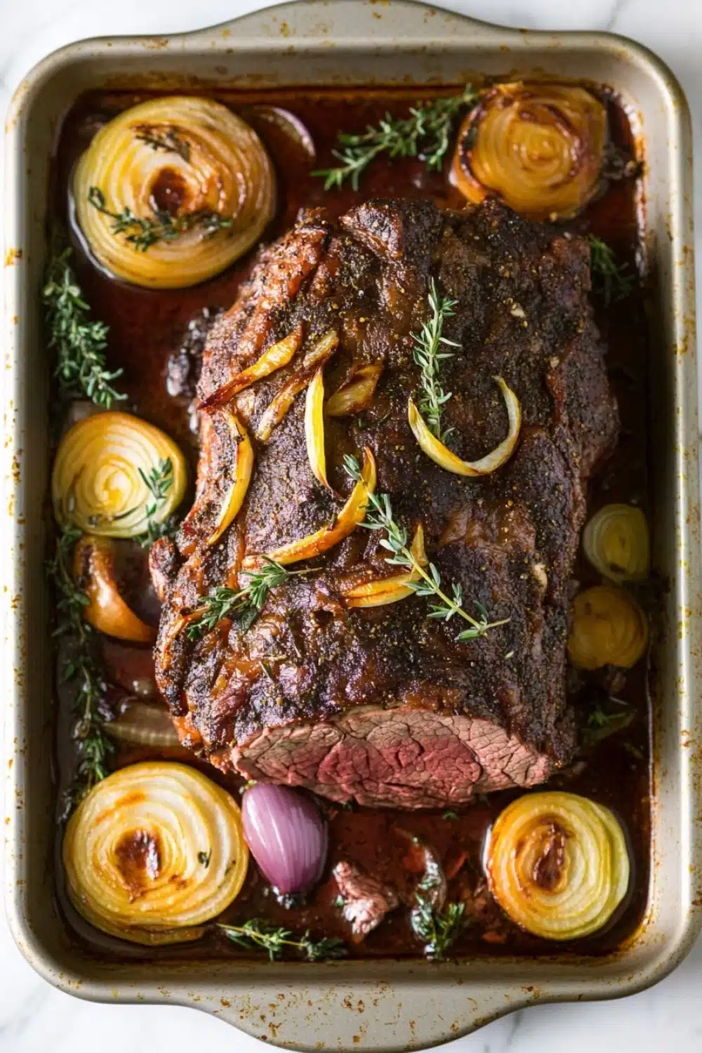 Keto slow cooker roast beef - the image shows a rectangular baking dish filled with a cooked steak. the steak appears to be medium-rare and has a dark brown crust on top. it is garnished with sliced onions and a sprig of thyme. the dish is sitting on a white marble countertop. there are also a few red onions scattered around the dish. the overall appearance of the dish is savory and appetizing.