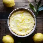 Lemon cake lemon pudding - the image is a top view of a lemon tart in a white ceramic bowl on a wooden table. the tart has a golden brown crust and is topped with a creamy yellow filling. there are three whole lemons on the table next to the tart, and a few green leaves scattered around the bowl. the background is dark, making the tart stand out. the overall mood of the image is fresh and appetizing.