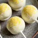 Lemon cake pop design - the image shows a plate with six cake pops on it. the cake pops are round and have a white base with yellow sprinkles on top. they are arranged in a circular pattern on the plate. the plate is brown and has a textured surface. the background is blurred, but it appears to be a kitchen countertop.