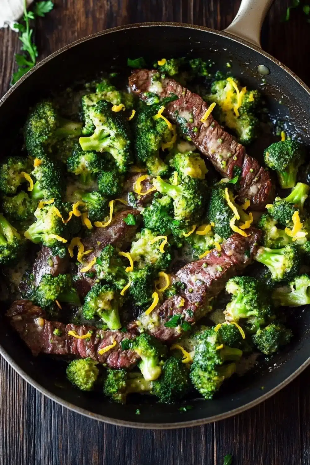 Lemon garlic butter steak and broccoli skillet - the image shows a black frying pan with a serving of cooked beef and broccoli in it. the beef appears to be medium-rare and has a pinkish-brown color. the broccoli is bright green and looks fresh and healthy. the pan is sitting on a wooden table with a few sprigs of parsley scattered around it. there is also a white napkin on the side of the pan. the overall appearance of the dish is savory and appetizing.