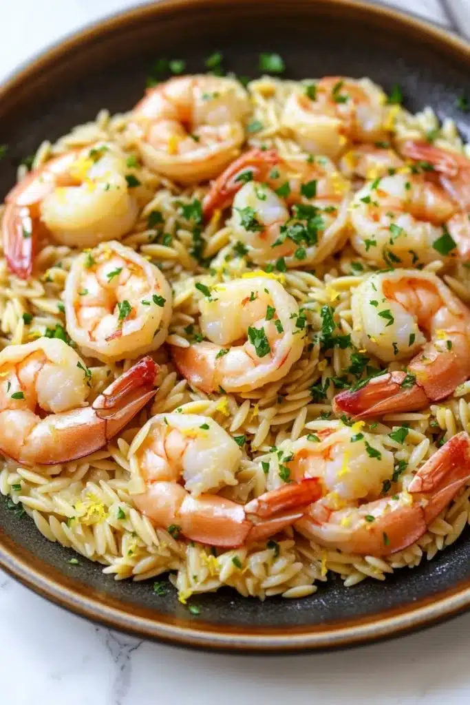 Lemon garlic shrimp orzo skillet - the image shows a plate of pasta dish with shrimp and lemon zest. the dish is served in a black bowl with a white marble countertop. the pasta appears to be fusilli, which is a type of spiral-shaped pasta, and is garnished with chopped parsley. the shrimp are cooked medium-sized and pink in color, and they are arranged in a circular pattern on top of the pasta. the parsley is a bright green color and is scattered throughout the dish. the overall appearance of the dish is fresh and appetizing.