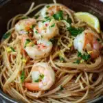 Low calorie shrimp scampi pasta - the image is a close-up of a bowl of spaghetti with shrimp and lemon wedges. the spaghetti is cooked al dente and is garnished with fresh parsley. the shrimp are large and pink, and they are arranged in a circular pattern on top of the spaghetti. the dish is served in a dark brown bowl with a wooden rim. the background is blurred, but it appears to be a rustic wooden table.