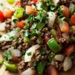 Low carb ground beef for dinner - the image shows a close-up of a dish on a wooden cutting board. the dish appears to be a mixture of ground beef, diced tomatoes, green bell peppers, onions, and cilantro. the ground beef is dark brown and looks to be seasoned with herbs and spices. the tomatoes are bright red and the onions are white. the cilantro is scattered throughout the dish, adding a pop of green color to the dish. the cutting board is a light-colored wood, and the background is blurred, making the dish the focal point of the image.