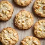 Macadamia nut shortbread cookie - the image shows a group of freshly baked cookies on a wooden surface. the cookies are round and golden brown in color, with a crumbly texture. they are arranged in a scattered manner, with some overlapping each other. some of the cookies have a bite taken out of one of them, revealing the white chocolate chips inside. the background is blurred, but it appears to be a rustic wooden table.