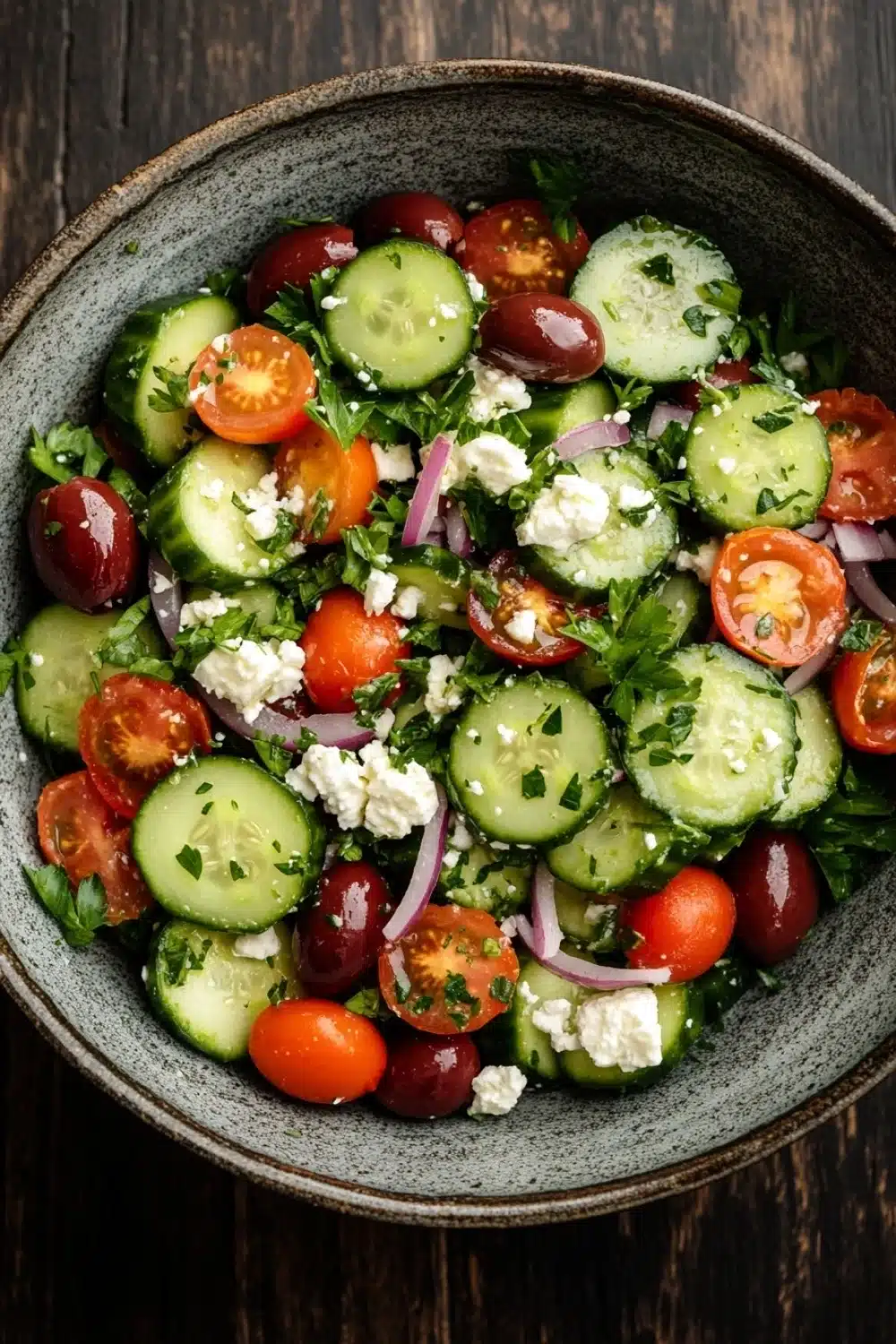 Mediterranean salad cucumber - the image is a close-up of a salad in a gray bowl. the salad is made up of sliced cucumbers, cherry tomatoes, red onions, and feta cheese. the cucumbers are arranged in a circular pattern, with some overlapping each other. the tomatoes are bright red and appear to be ripe, while the onions are sliced into thin strips. the feta is crumbled and is scattered throughout the salad. there are also some green herbs, possibly parsley, adding a pop of color to the dish. the bowl is sitting on a dark wooden table.