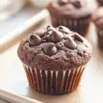 Muffin cacao e yogurt - the image shows a close-up of a freshly baked chocolate muffin on a white baking tray. the muffin is golden brown in color and has a crumbly texture. it is topped with chocolate chips, giving it a shiny appearance. in the background, there are more muffins and a white cloth napkin. the tray is lined with parchment paper, and the background is blurred, making the muffins the focal point of the image.