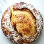 No knead dutch oven bread - the image is a close-up of a round loaf of bread on a white marble surface. the bread appears to be freshly baked and has a golden brown crust on top. the inside of the bread is visible, with a layer of melted cheese oozing out of it. the cheese is a light yellow color and looks soft and fluffy. the crust is slightly crinkled, giving the bread a textured appearance. the overall color of the image is warm and inviting, with shades of brown, white, and gray.