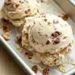 Oatmeal cookie ice cream - the image shows three scoops of vanilla ice cream in a rectangular baking tray. the ice cream is a light beige color and has a crumbly texture. it is topped with chopped pecans, giving it a crunchy appearance. the tray is sitting on a wooden table with a spoon resting on the side. the background is blurred, but it appears to be a kitchen countertop.