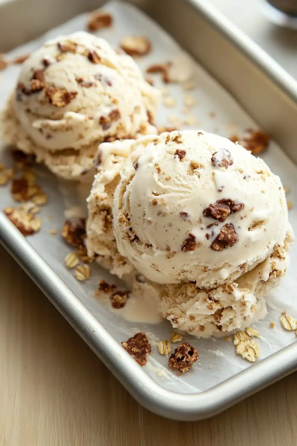 Oatmeal cookie ice cream - the image shows three scoops of vanilla ice cream in a rectangular baking tray. the ice cream is a light beige color and has a crumbly texture. it is topped with chopped pecans, giving it a crunchy appearance. the tray is sitting on a wooden table with a spoon resting on the side. the background is blurred, but it appears to be a kitchen countertop.