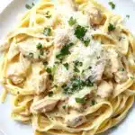 One pot chicken fettuccine - the image shows a plate of fettuccine alfredo pasta with chunks of chicken and parmesan cheese on top. the pasta is cooked al dente and is covered in a creamy white sauce. the dish is garnished with fresh parsley leaves. the plate is white and is sitting on a white marble countertop. the overall presentation of the dish is elegant and appetizing.